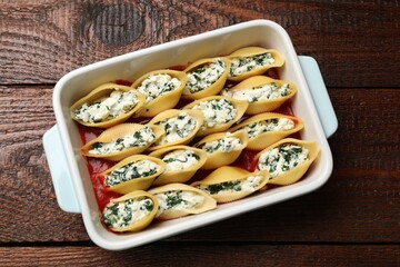 Delicious conchiglie pasta with ricotta cheese, spinach and tomato sauce in baking dish on wooden table, top view