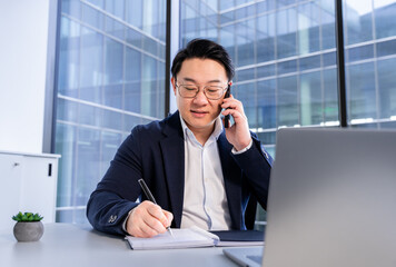 Confident businessman in formal attire discussing over phone call while taking notes in a notepad....