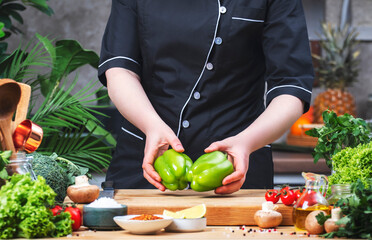 Chef holding two green paprika in hands. Cozy kitchen with wooden table, kitchenware, vegetables, herbs and ingredients for cooking. Healthy vegan eating, culinary, recipes, food blogging