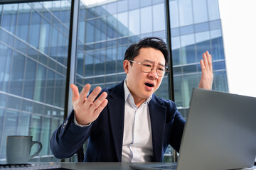 Fototapeta premium Asian businessman sitting at a desk in a modern office, facing a laptop, appearing frustrated and expressive. His corporate attire conveys professional circumstances and modern workplace culture.