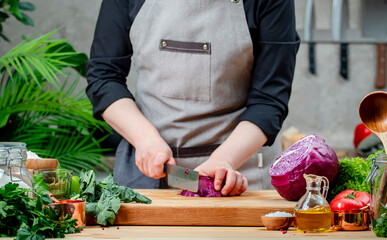 Chef cuts organic ​​red cabbage. Cozy kitchen with wooden table, kitchenware, vegetables, herbs and ingredients for cooking. Healthy vegan eating, culinary, recipes, food blogging comcept