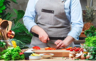 Chef cuts red paprika or bell pepper. Cozy kitchen with wooden table, kitchenware, vegetables, herbs and ingredients for cooking. Healthy vegan eating, culinary, recipes, food blogging