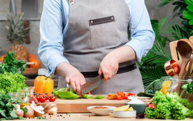 Chef cuts green paprika or bell pepper. Cozy kitchen with wooden table, kitchenware, vegetables, herbs and ingredients for cooking. Healthy vegan eating, culinary, recipes, food blogging
