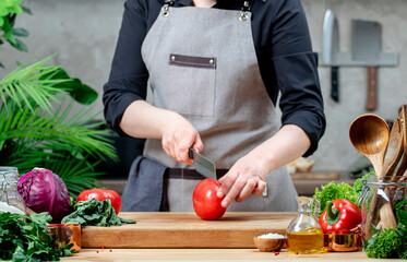 Chef cuts in half big red tomato. Cozy kitchen with wooden table, kitchenware, vegetables, herbs and ingredients for cooking. Healthy vegan food, culinary, recipes, food blogging