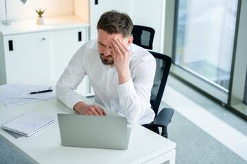 Fototapeta premium Professional businessman sits in a modern office, appearing stressed while working on a laptop. Papers and notebooks surround him, portraying a stressful and busy work environment. Business concept.