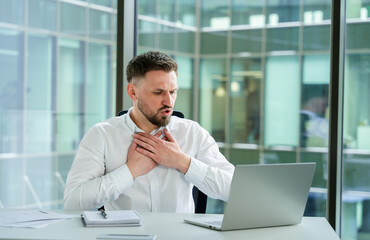 Sick businessman in modern office holding chest, working on laptop, feeling worried and distracted....