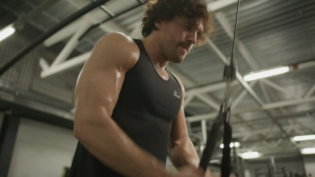 Strong man with muscular body working out on pulley machine in gym, performing triceps pushdown exercise with effort. Low angle view