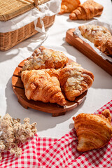 Golden flaky croissants with almond toppings displayed on a rustic wooden board, set on a white tablecloth with a red checkered napkin, creating a cozy and inviting bakery atmosphere