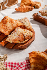 Golden flaky croissants with almond toppings displayed on a rustic wooden board, set on a white tablecloth with a red checkered napkin, creating a cozy and inviting bakery atmosphere