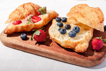 Delicious breakfast spread featuring buttery croissants topped with fresh strawberries and blueberries, served on a wooden board with butter, coffee, and fruit, creating a cozy