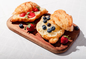 Delicious breakfast spread featuring buttery croissants topped with fresh strawberries and blueberries, served on a wooden board with butter, coffee, and fruit, creating a cozy