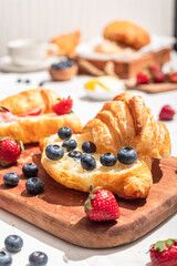 Delicious breakfast spread featuring buttery croissants topped with fresh strawberries and blueberries, served on a wooden board with butter, coffee, and fruit, creating a cozy