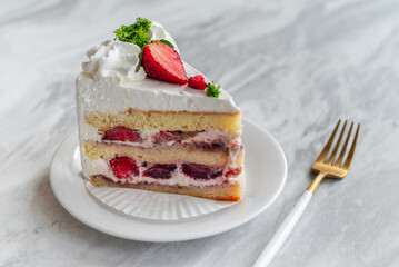 Slice of strawberry shortcake with layers of cream and fresh strawberries, garnished with whipped cream and parsley, served on a white plate with a gold fork on a marble table