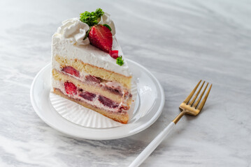Slice of strawberry shortcake with layers of cream and fresh strawberries, garnished with whipped cream and parsley, served on a white plate with a gold fork on a marble table