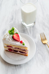 Slice of strawberry shortcake with whipped cream and fresh strawberry garnish on a white plate, accompanied by a glass of milk and a gold fork on a marble table, ideal for dessert lovers