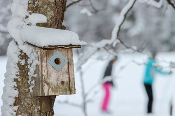 An empty birdhouse covered in snow awaits spring and new residents. In a blurred background there are two skier figures dressed in colorful clothes.