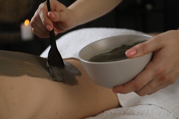 Esthetician applying cosmetic product for body wraps treatment onto woman's back in spa salon, closeup