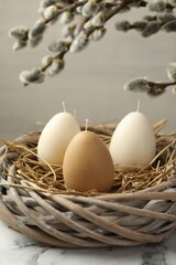 Egg-shaped candles in nest on white marble table and willow branches against light background, closeup. Easter decor