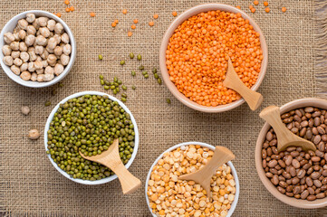 An assortment of legume products: chickpeas, mung beans, lentils, peas, beans in bowls on a cloth, burlap. Top view, horizontal. Healthy nutrition, vegetarianism.