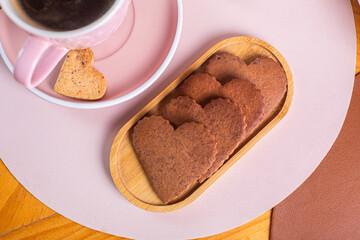 Heart-shaped cookies on a wooden plate beside a cup of coffee on a pink table, perfect for a cozy afternoon snack, sweet treat concept