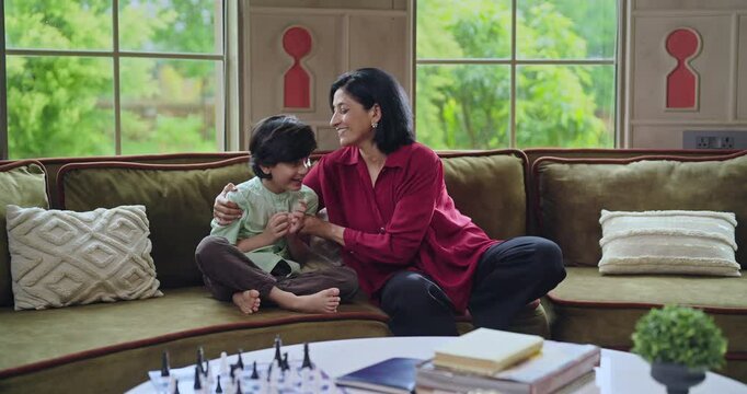 Indian happy adult mother cute kid son sitting on cozy sofa couch talk gossip do tickle have fun joy young lady mom boy child play casual game laugh enjoy good day spend time feel free indoor home	