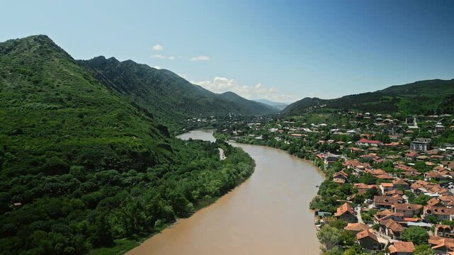 Aerial view of the Mtkvari River winding through a lush green valley near Tbilisi, Georgia. Traditional houses with red roofs contrast against the mountainous landscape