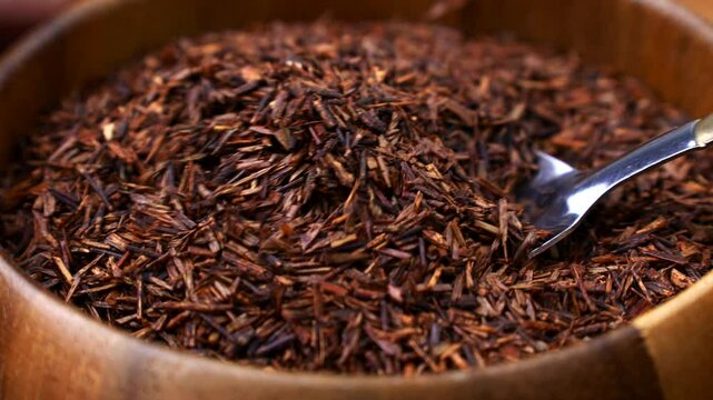 Rooibos tea dried leaves taking with a spoon from a wooden bowl close up