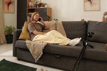 Obraz premium Woman sitting on couch while talking on phone and smiling warmly. Sofa with pillows, nearby shelves with books and plant add homely touch