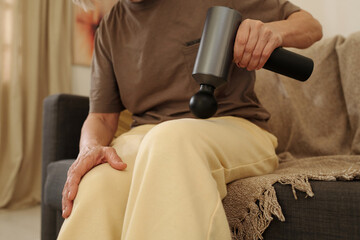 Fototapeta premium Individual sitting on couch using percussive massage gun on their leg for muscle relief, enhancing relaxation and recovery experience in home environment