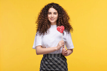 Cheerful curly-haired woman holding heart-shaped lollipop on yellow background, expressing joy and...