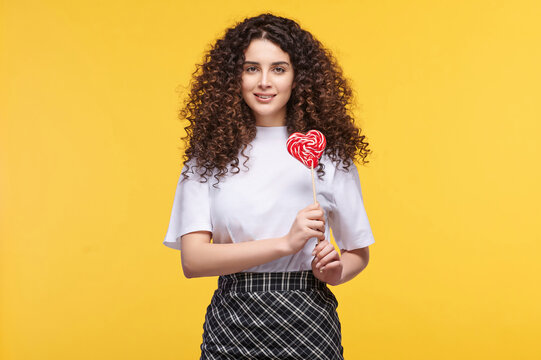 Cheerful curly-haired woman holding heart-shaped lollipop on yellow background, expressing joy and playfulness