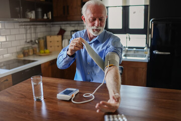 An elderly man with grey hair and a beard sits in his home kitchen, carefully putting on a digital...