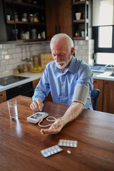 An elderly man with grey hair and a beard sits in his home kitchen, using a digital blood pressure...