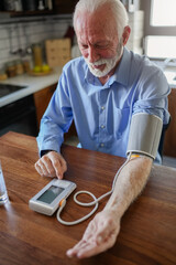 An elderly man with grey hair and a beard sits in his home kitchen, using a digital blood pressure...