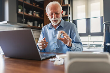 Fototapeta premium Elderly man in his home kitchen with a laptop in front of him and various pills on the table carefully examines a plastic pill container, reading the label, possibly verifying dosage or instructions