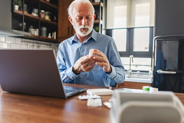 Elderly man in his home kitchen with a laptop in front of him and various pills on the table...