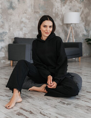 Portrait of a cute girl in a black hoodie and black jeans on the background of a sofa in a room sitting on the floor