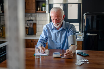 An elderly man with grey hair and a beard sits in his home kitchen, measuring his blood pressure...