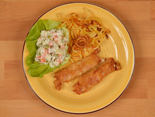 An overhead shot of a yellow plate featuring two fried rolls, a side of potato salad, and fried potato noodles on a wooden table, showcasing a simple yet satisfying meal