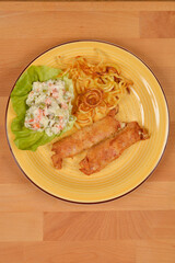 An overhead shot of a yellow plate featuring two fried rolls, a side of potato salad, and fried potato noodles on a wooden table, showcasing a simple yet satisfying meal