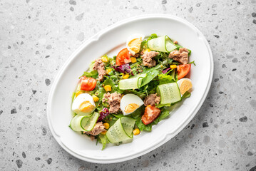 A fresh and vibrant salad featuring mixed greens, sliced cucumbers, cherry tomatoes, tuna, corn, and boiled eggs, elegantly presented on a white oval plate