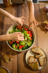 Small children’s hands reach for fresh vegetables from a shared salad bowl on the table. A colorful and healthy meal encourages kids to enjoy fresh food together.