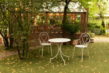 Fototapeta premium White iron chairs and table at outdoor or outside for relax. Autumn atmosphere in garden - table and chairs and fallen yellow leaves. Backyard with garden furniture set. Interior cozy patio at home. 