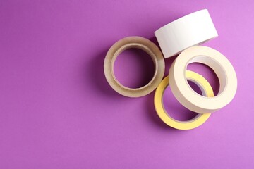 Rolls of different adhesive tapes on purple background, flat lay. Space for text