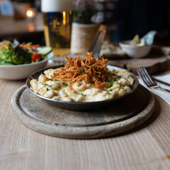 Herzhafte Käsespätzle mit Röstzwiebeln in Gusseisenpfanne, serviert im gemütlichen Wirtshaus mit frischem Salat und kühlem Bier, bayerische Alpenküche

