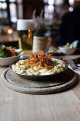 Cremige Käsespätzle mit knusprigen Röstzwiebeln in Gusseisenpfanne, serviert mit frischem Salat und Bier im rustikalen Wirtshaus, gemütliche Pub-Atmosphäre, traditionelle deutsche Küche


