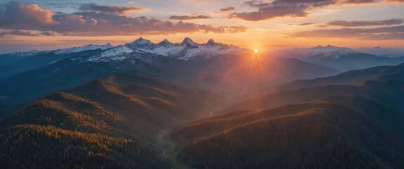 Aerial view of mountain landscape during sunrise with snow-capped peaks and golden sunlight illuminating forested hills in the foreground