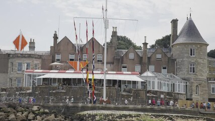 A group of people are sitting on a pier near a large building. The pier is surrounded by a lot of flags, and there is a boat in the water. Scene is peaceful and relaxing - Powered by Adobe