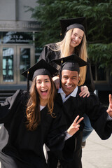 Three cheerful multi ethnic graduates wearing gowns and mortarboards gesturing victory sign...