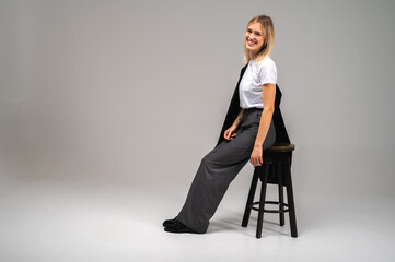 Smiling young woman in stylish business casual outfit sitting on stool on neutral background....
